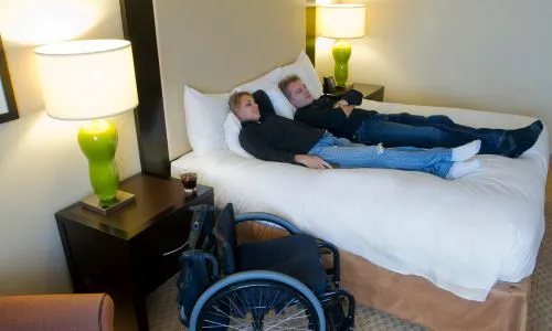 Deux personnes dans un lit près d’un fauteuil roulant.