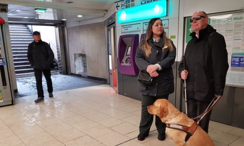 Un homme aveugle tenant son chien guide parle avec un agent RATP dans 1 station.