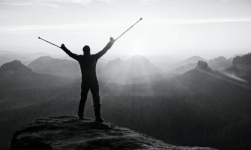 Un homme avec des béquilles, en haut d’une montagne, lève les bras vers le ciel.