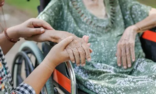 Une jeune femme tient la main d’une personne âgée en fauteuil roulant.