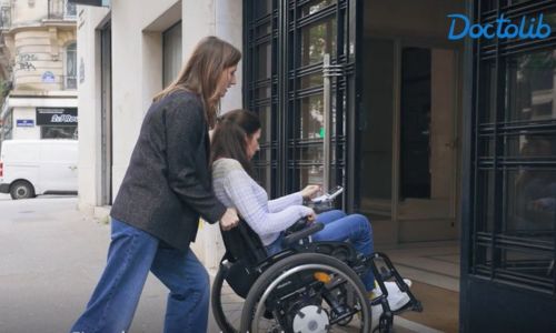 Virginie Dubost, en fauteuil roulant, aidée par une femme pour gravir une marche