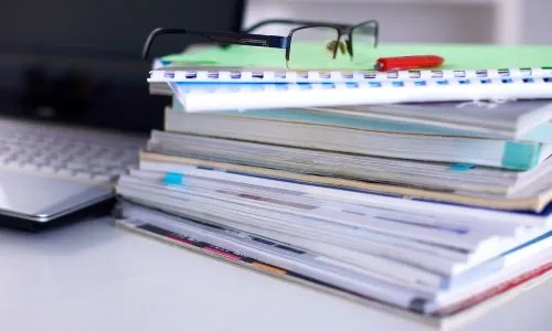 Des lunettes posées sur une pile de dossiers, près d’un ordinateur.