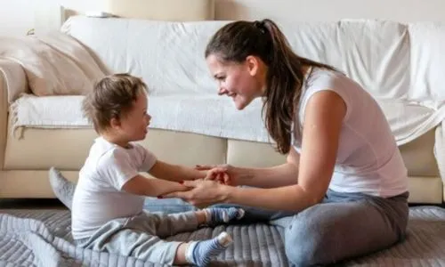 Une maman assise sur un tapis en face de son enfant à qui elle tient les mains.