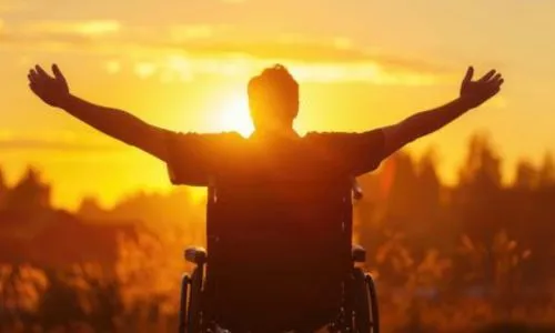 Un homme en fauteuil lève les bras, surplombant une ville au coucher du soleil.