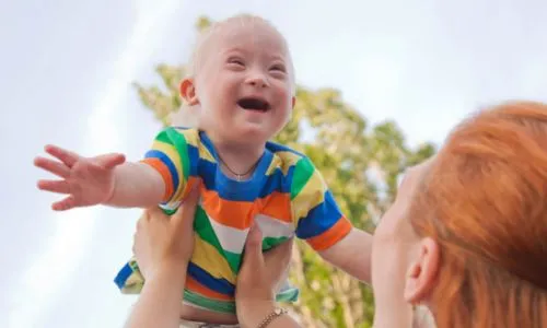 Une maman porte son bébé avec trisomie 21 dans les airs.